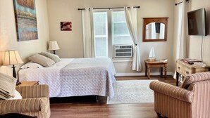 Un dormitorio con una cama, dos sillas, una mesita, un ventilador de techo, una ventana con cortinas, un espejo y un televisor montado en la pared.