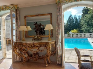 An ornate gold console table with a mirror and two lamps, a pool in the background, and a view of trees and a white fence. - DORDOGNE VACATION RENTALS (Thiviers)