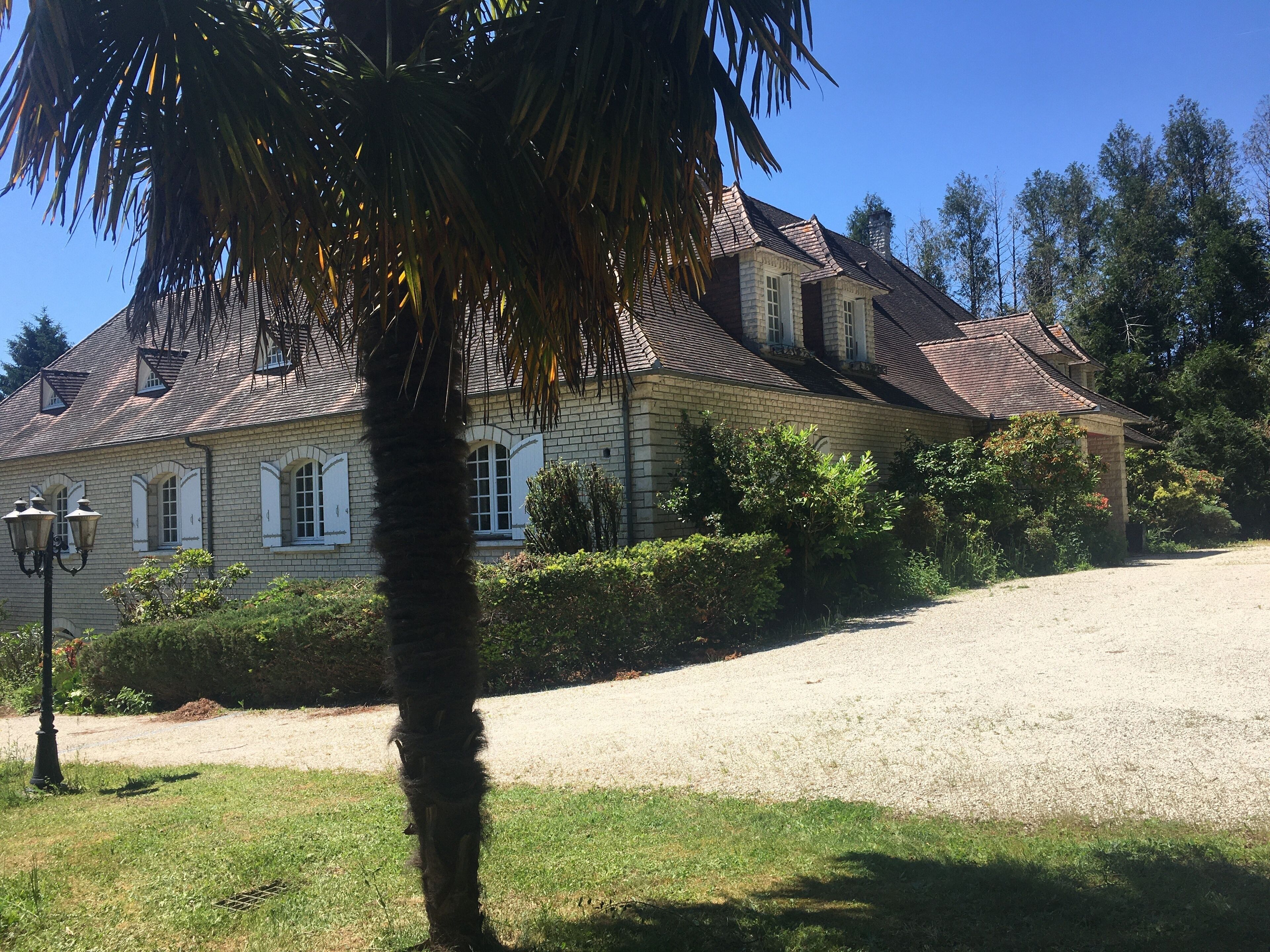 Une grande maison à deux étages, avec une façade en briques, de nombreuses fenêtres dotées de volets et une allée gravillonnée.