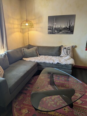 Una sala de estar con un sofá gris, una mesa de centro de vidrio, un tapete estampado, un cuadro colgado en la pared y una lámpara colgante.