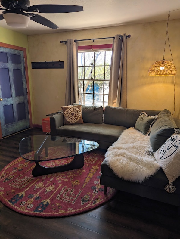 Una sala de estar con un sofá, una mesa de centro de vidrio, un tapete estampado y una ventana con cortinas.