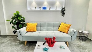 A modern living room with a grey sofa, yellow cushions, a white coffee table with books and decorative items, a large green plant, and a wall-mounted shelf with various objects. - Il Rifugio (Marettimo)