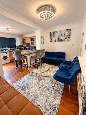 A modern living room with a blue sofa, a round coffee table, and a chandelier. - Modern Home for Contractors - Sleeps 10+Driveway - Near Central London & Airport (Barking)