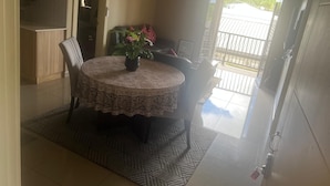 A dining area with a round table, chairs, and a potted plant. There is a door leading to a balcony with a view of the outside.