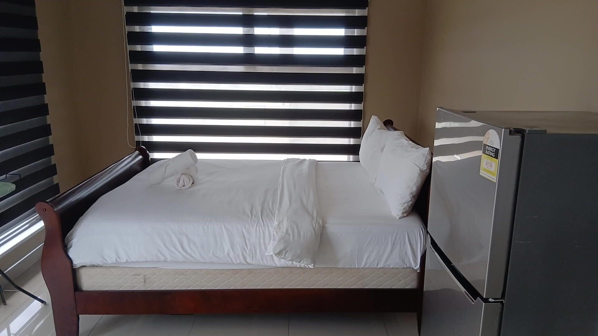 Un lit simple avec du linge de lit blanc, un cadre en bois et un oreiller blanc. Un réfrigérateur, muni d’une étiquette indiquant sa classe énergétique, se trouve à droite du lit.