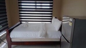 A single bed with white linens, a wooden frame, and a white pillow. There is a refrigerator with an energy rating sticker to the right of the bed.