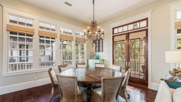 A dining room with a round wooden table surrounded by wicker chairs, large windows with blinds, and a chandelier. - Community Pool Access with Carriage (Rosemary Beach)