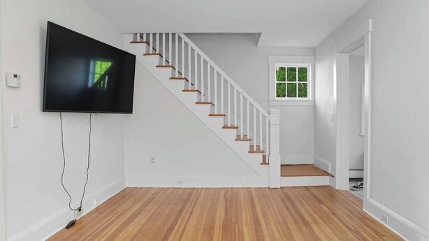 Ein geräumiges Zimmer mit Holzboden, einer weißen Treppe, einem an der Wand montierten Fernseher und einem Fenster mit Blick auf Grünflächen.