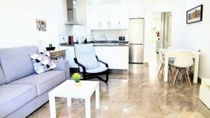 Een moderne woonkamer met een grijze bank, een witte salontafel en een zwarte fauteuil. Er is een keukenhoek met witte kasten en een roestvrijstalen koelkast.
