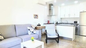Een moderne woonkamer met een grijze bank, een witte salontafel en een klein bijzettafeltje met een vaas. Een keuken is zichtbaar met witte kasten en een roestvrijstalen koelkast.