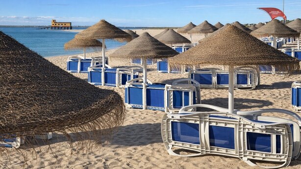Una playa con sombrillas de paja y sillones azules.