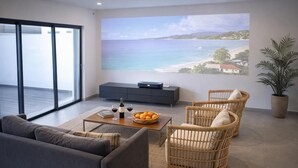 Una sala de estar moderna con un ventanal grande, un sofá, dos sillones de mimbre, una mesita de centro con una botella y vasos, y vista al mar.