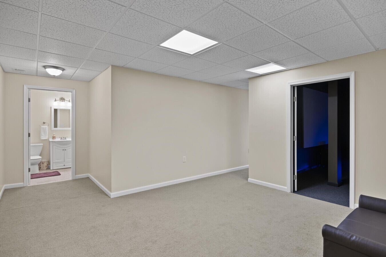 A spacious room with a carpeted floor, a door leading to a bathroom, and a dark room visible through an open door.