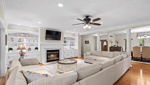 A spacious living room with a fireplace, a large sectional sofa, a ceiling fan, and a television mounted above the fireplace. - 8,000 Sq Ft Luxury Estate Near Annapolis | Sleeps 32 | Pool, Theater & Pickleball (Davidsonville)
