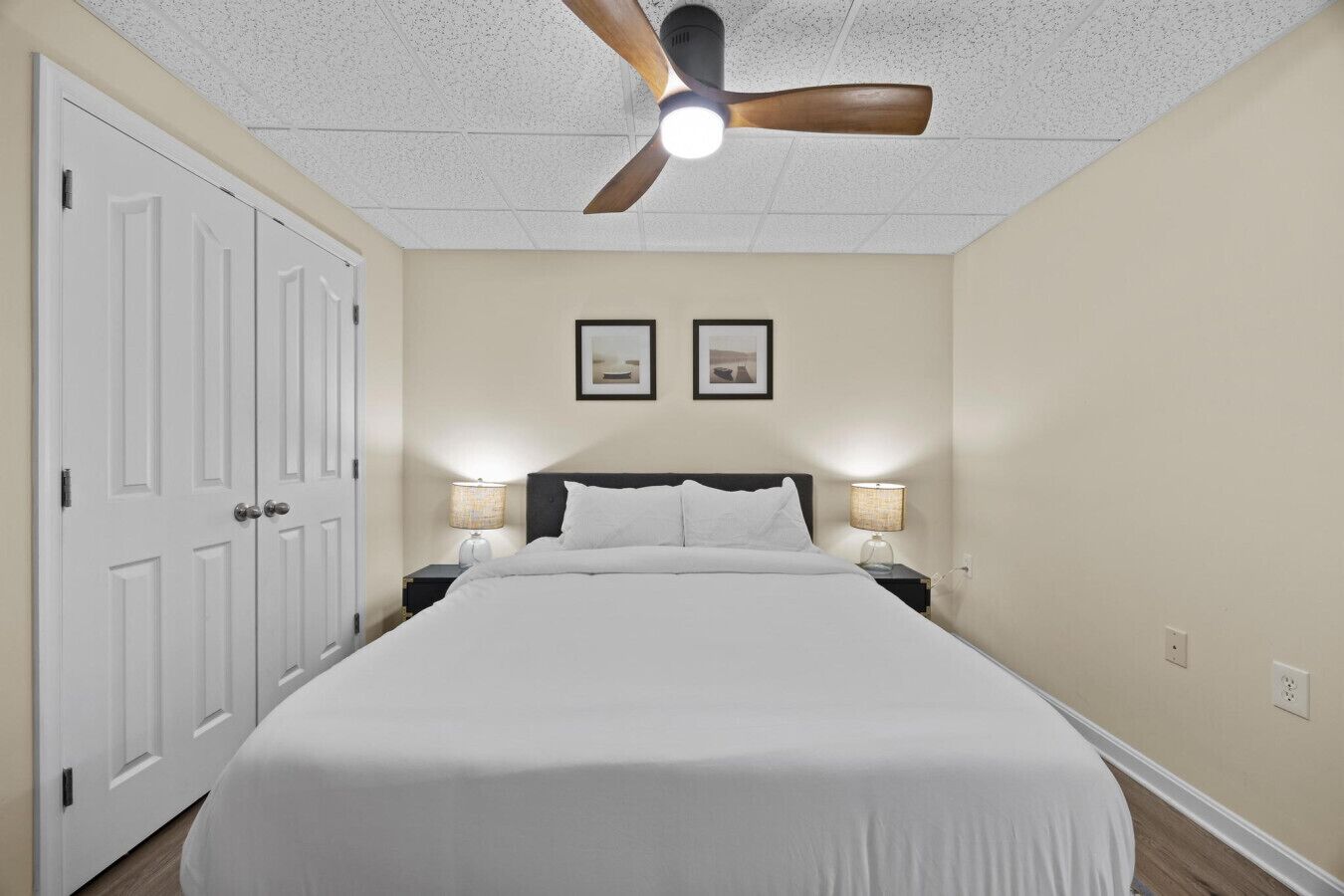 A bedroom with a bed, two bedside lamps, a ceiling fan, and two framed pictures on the wall.