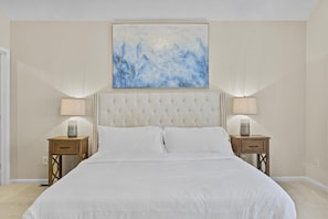 A bedroom with a tufted headboard, two bedside tables with lamps, a large abstract painting above the bed, and a neutral color scheme.