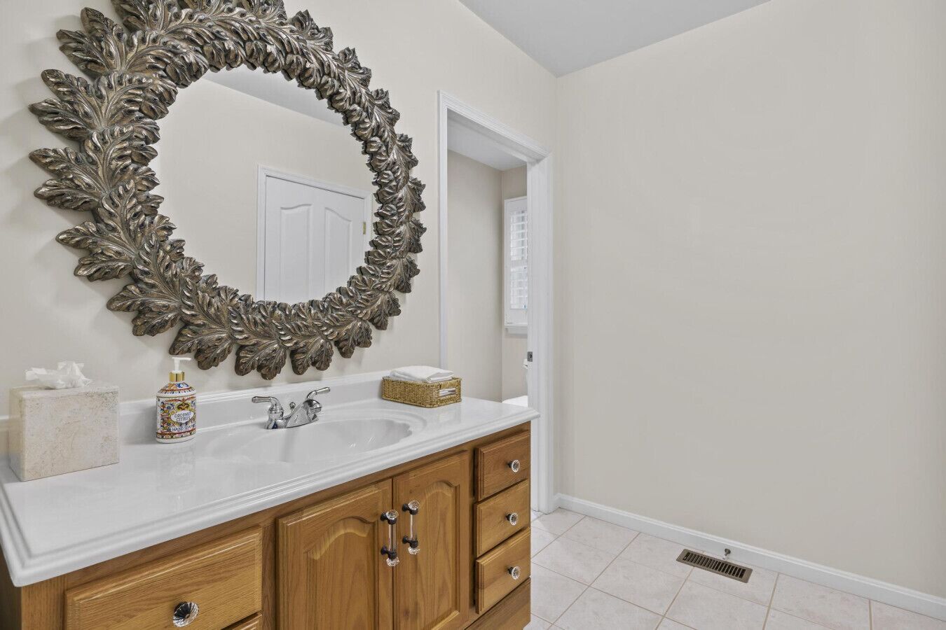 A bathroom with a large ornate mirror, a white sink, wooden cabinets, and a white door in the background.