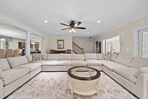 A living room with a sectional sofa, a coffee table, a ceiling fan, and a view into the kitchen.