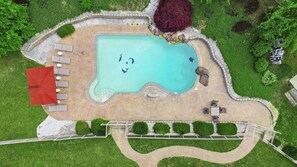 Luftaufnahme eines Swimmingpools mit einem roten Sonnenschirm, Liegestühlen und umgebender Bepflanzung.