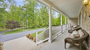 A covered porch with wicker furniture, a stone wall, and a view of a tree-lined street. - 8,000 Sq Ft Luxury Estate Near Annapolis | Sleeps 32 | Pool, Theater & Pickleball (Davidsonville)