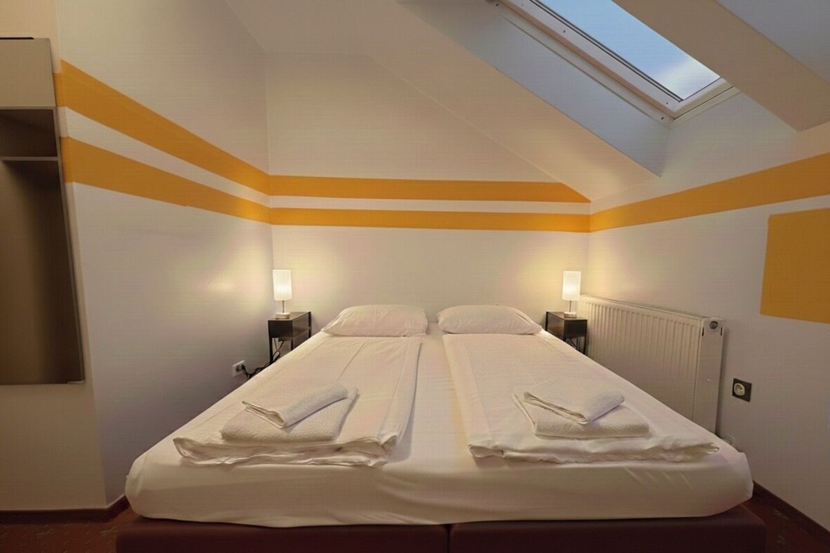 Una habitación de hotel con una cama doble, dos mesitas de noche con lámparas, un tragaluz y un radiador.