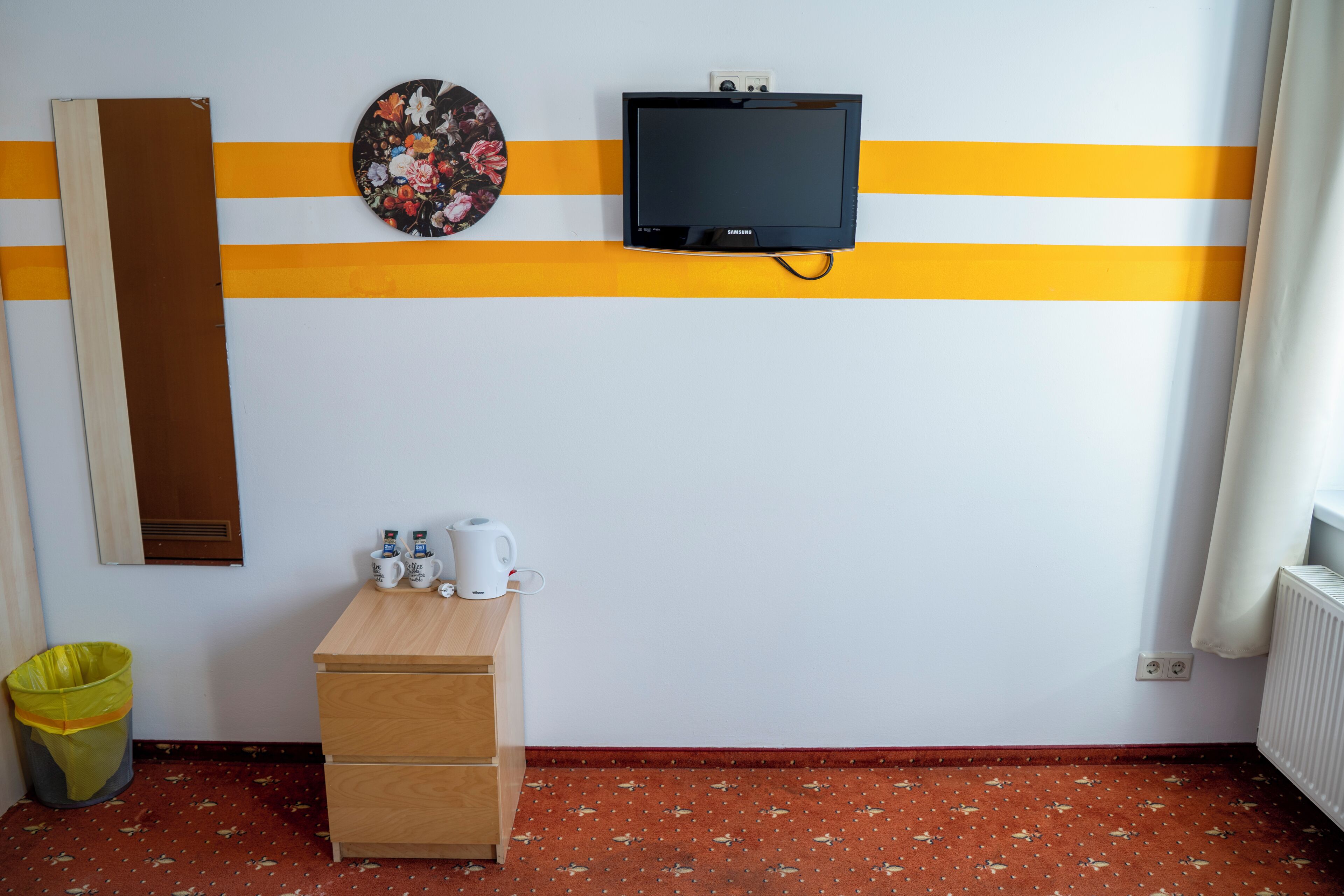 Ein Zimmer mit Holzschreibtisch, Mülleimer, Wandfernseher und Heizkörper.