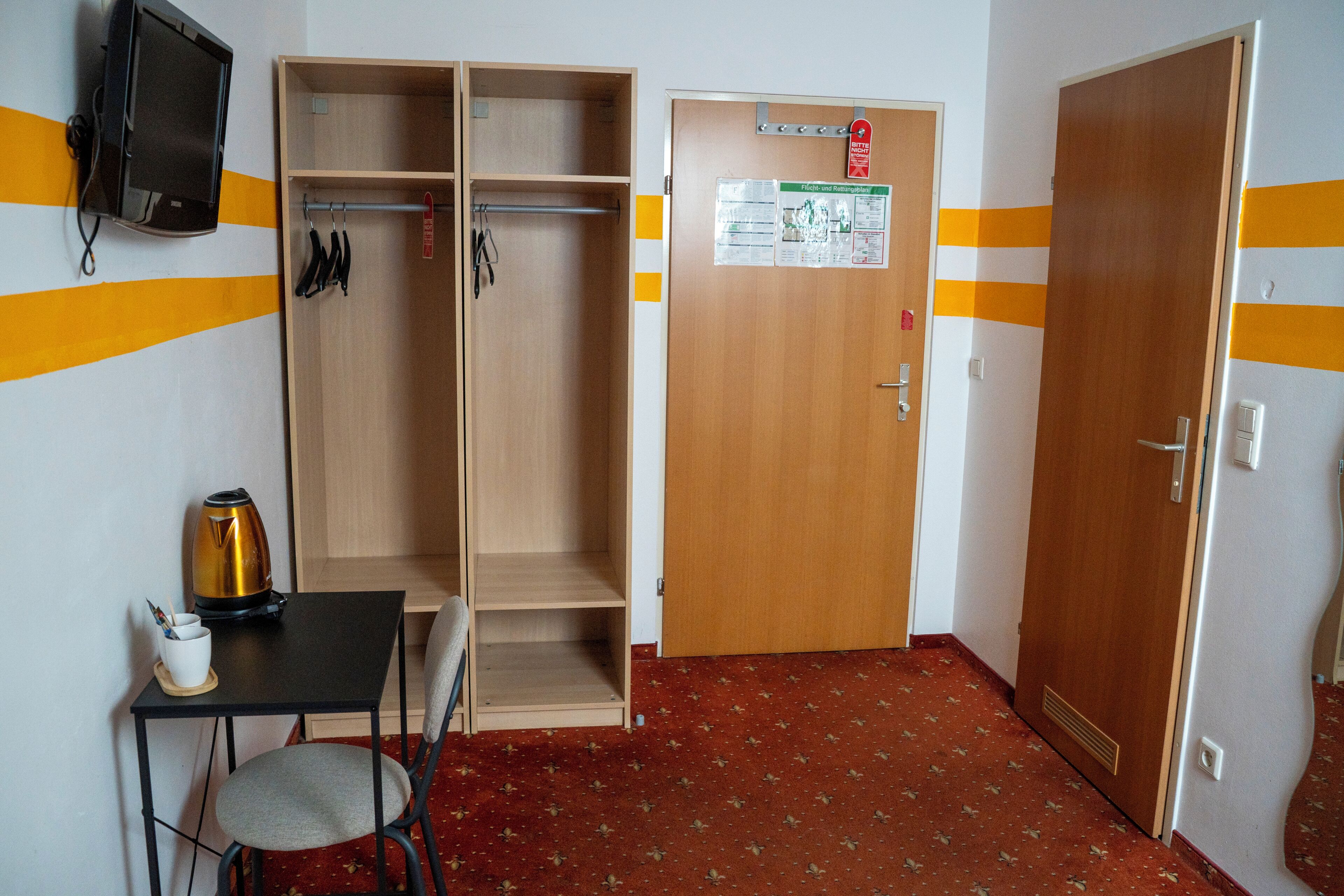 A room with a wooden wardrobe, a small table with a kettle, a chair, and a television mounted on the wall.
