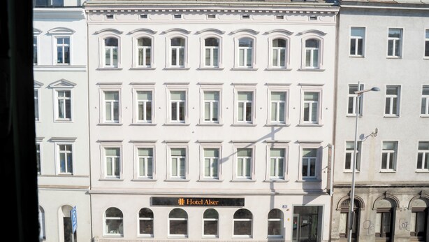 Un edificio de varios pisos con un estilo arquitectónico clásico, que presenta un llamativo letrero del 'Hotel Alsser' a nivel de calle.
