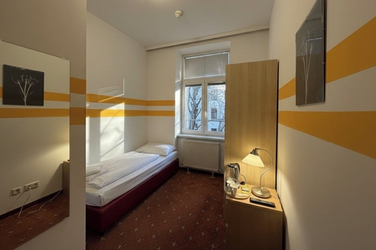 Una habitación de hotel con una cama individual, un radiador, una ventana con persianas, una mesita de noche con una lámpara y una pared con diseño de rayas amarillas.