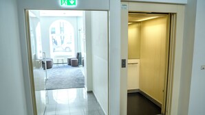 Un vestíbulo de hotel con un puerta de vidrio transparente que da a un espacio exterior, una recepción y una escalera hacia los ascensores.