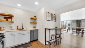 Une cuisine moderne avec des meubles blancs, des étagères en bois et un coin repas donnant sur l’extérieur grâce à une porte vitrée.