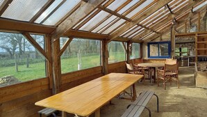 Ein rustikaler Essbereich aus Holz mit einem langen Tisch, Bänken, großen Fenstern und einem Kamin aus Stein.