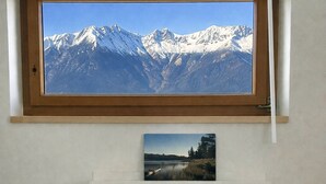 Habitación con un ventanal que muestra un paisaje montañoso, una fotografía más pequeña debajo y un cabecero de madera con almohadas.