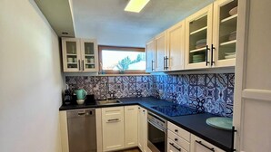 Cocina compacta con muebles blancos, lavavajillas, horno y ventana con vista a árboles.
