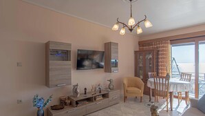 A living room with a TV, wooden cabinet, sofa, and dining area with a table and chairs. - Feliz Apt- Breathtaking Seaview and Sunset by ΜΡS (Anavissos)