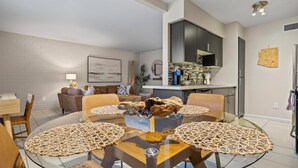 A modern living room with a glass coffee table, leather sofas, and a kitchen area with dark cabinets and a backsplash.