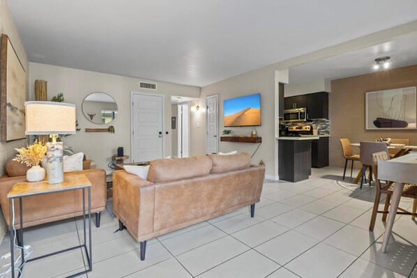 A modern living room with a sofa, a coffee table, and a TV. There is a kitchen area with a microwave and a dining table with chairs.