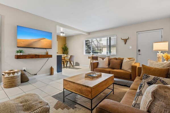 A modern living room with a large TV, a sofa, a coffee table, and a dining area with a staircase visible in the background.