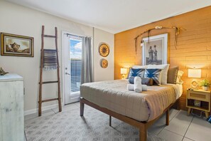 A bedroom with a wooden bed, a ladder, a window with curtains, a wall clock, and a small bedside table with a lamp.