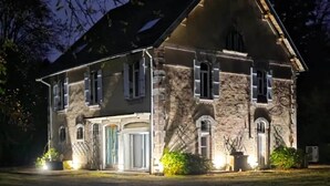 Un edificio de piedra con tejado a dos aguas y varias ventanas, iluminado por luces exteriores.