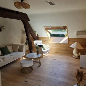 A cozy loft-style room with a wooden floor, a sofa, a chair, a small table, and a window with a view of greenery. - Chalet des Loups - "Le refuge" luxury wellness suite (Breuches)
