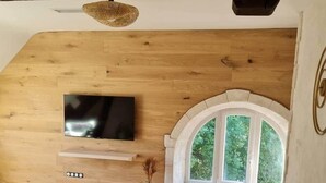 Una habitación acogedora con techo de madera, un televisor de pantalla plana en la pared, una ventana en arco de piedra y un cofre de madera rústica.