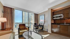 A hotel room with a large window, a dining table, a chair, a sofa, a glass coffee table, a lamp, a microwave, and a sink. - Luxurious Tower 3 Studio | Pool + Strip View (Las Vegas)