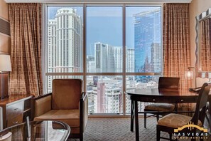A hotel room with a large window, a dining table, two chairs, a sofa, and a lamp.