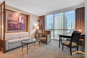 A hotel room with a sofa, armchair, dining table, and a large window with a city view.