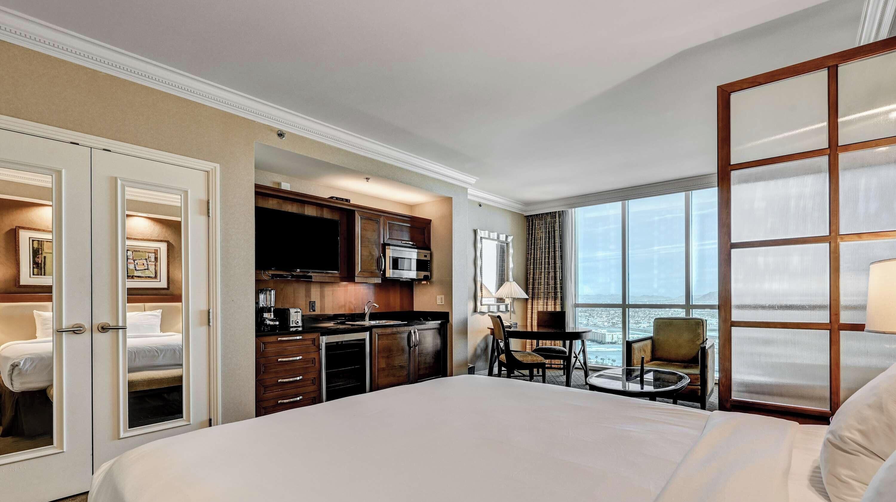 A hotel room with a bed, a kitchenette, a dining area, and a large window with a view.