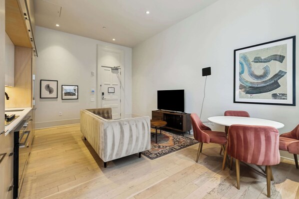 Ein modernes Wohnzimmer mit einem weißen Sofa, einem runden Tisch und rosafarbenen Stühlen. Ein Flachbildfernseher und ein großes abstraktes Gemälde sind an der Wand angebracht.
