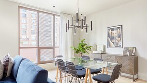 A modern living room with a glass dining table, black chairs, a blue sofa, and a large window with a view of buildings. - Sleek 2 BR Apartment | Near Bourbon St. + Superdome (New Orleans)