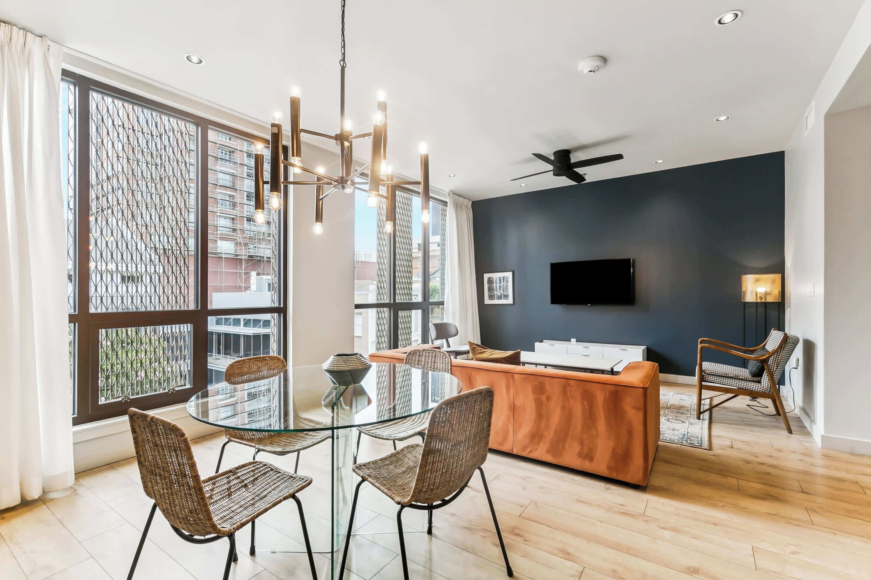 A modern living room with a glass dining table, wicker chairs, a wooden sofa, a TV mounted on the wall, and large windows with a view of buildings.