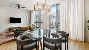 Ein modernes Wohnzimmer mit einem Glastisch, bequemen Sitzmöglichkeiten, einem Flachbildfernseher und großen Fenstern mit Vorhängen.